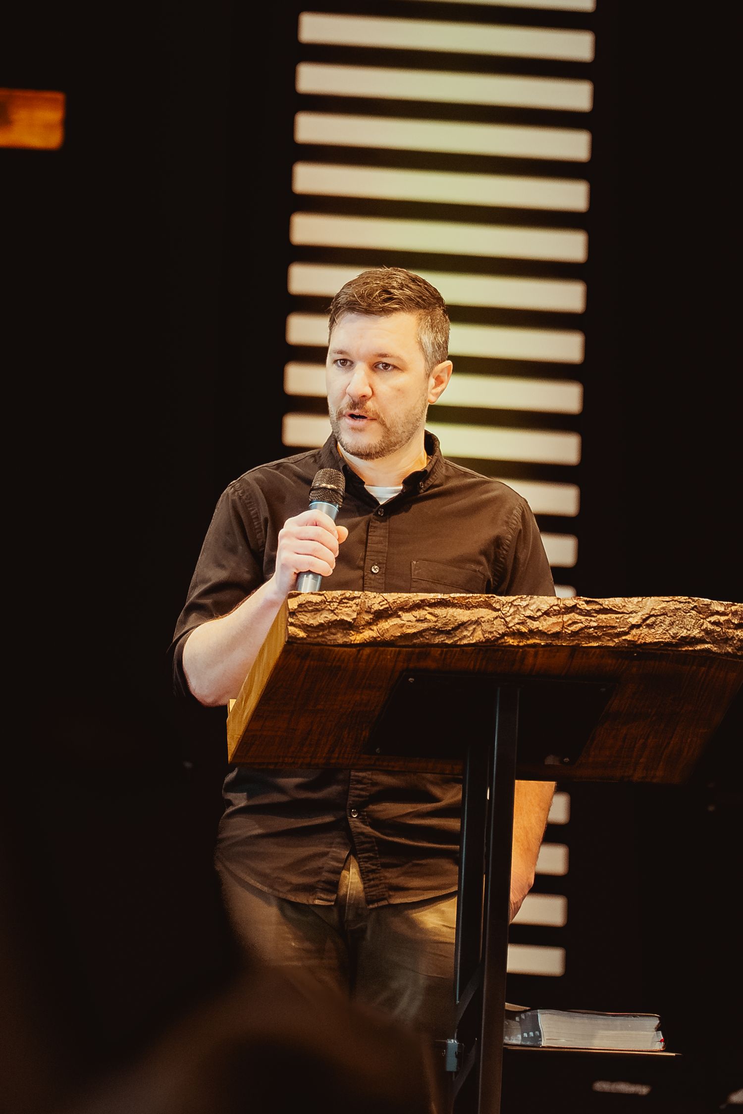 A speaker in a brown shirt sits at a dark podium on stage with horizontal white stripes in the background.
