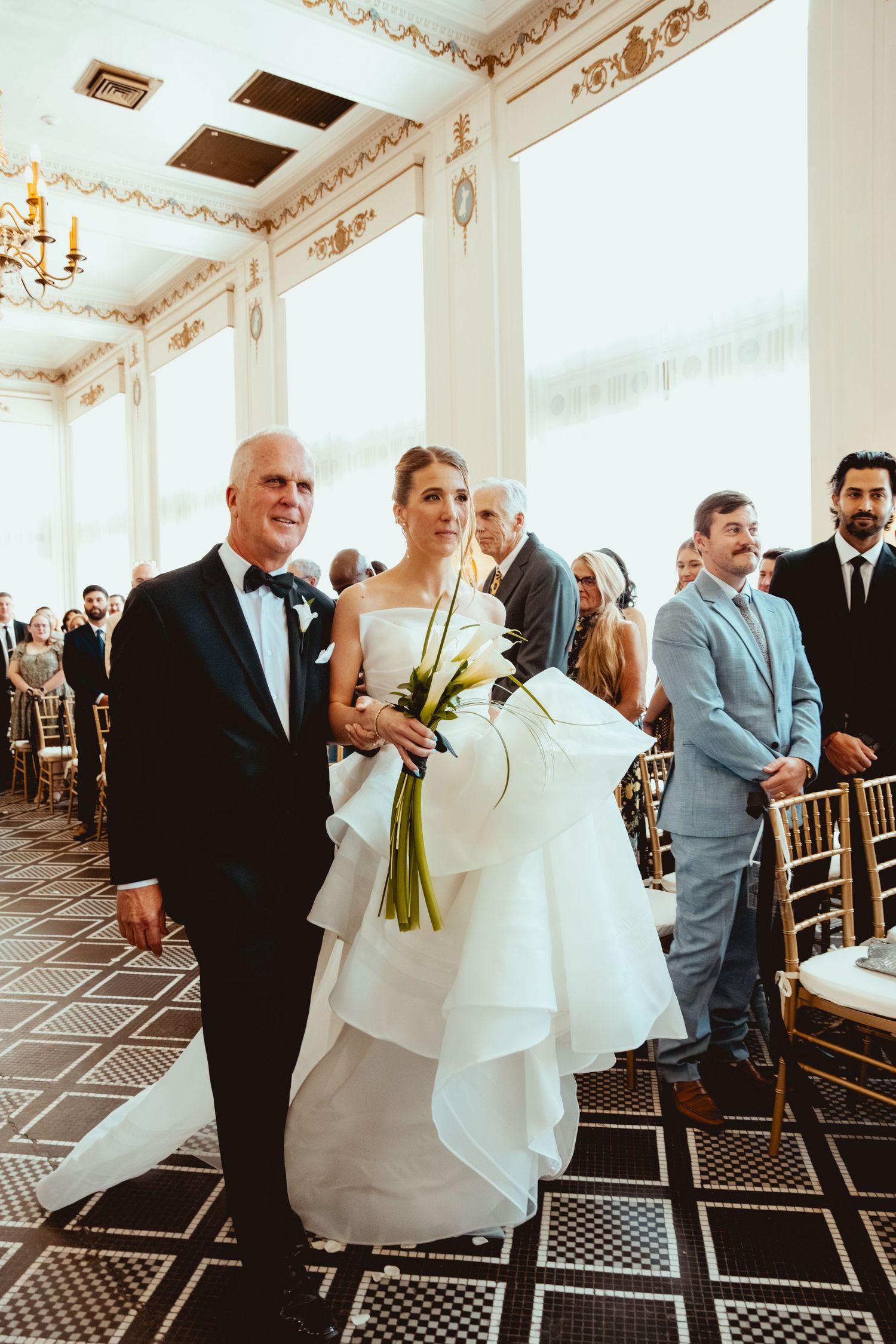 Wedding ceremony guests stand in a grand white ballroom with geometric patterned flooring.