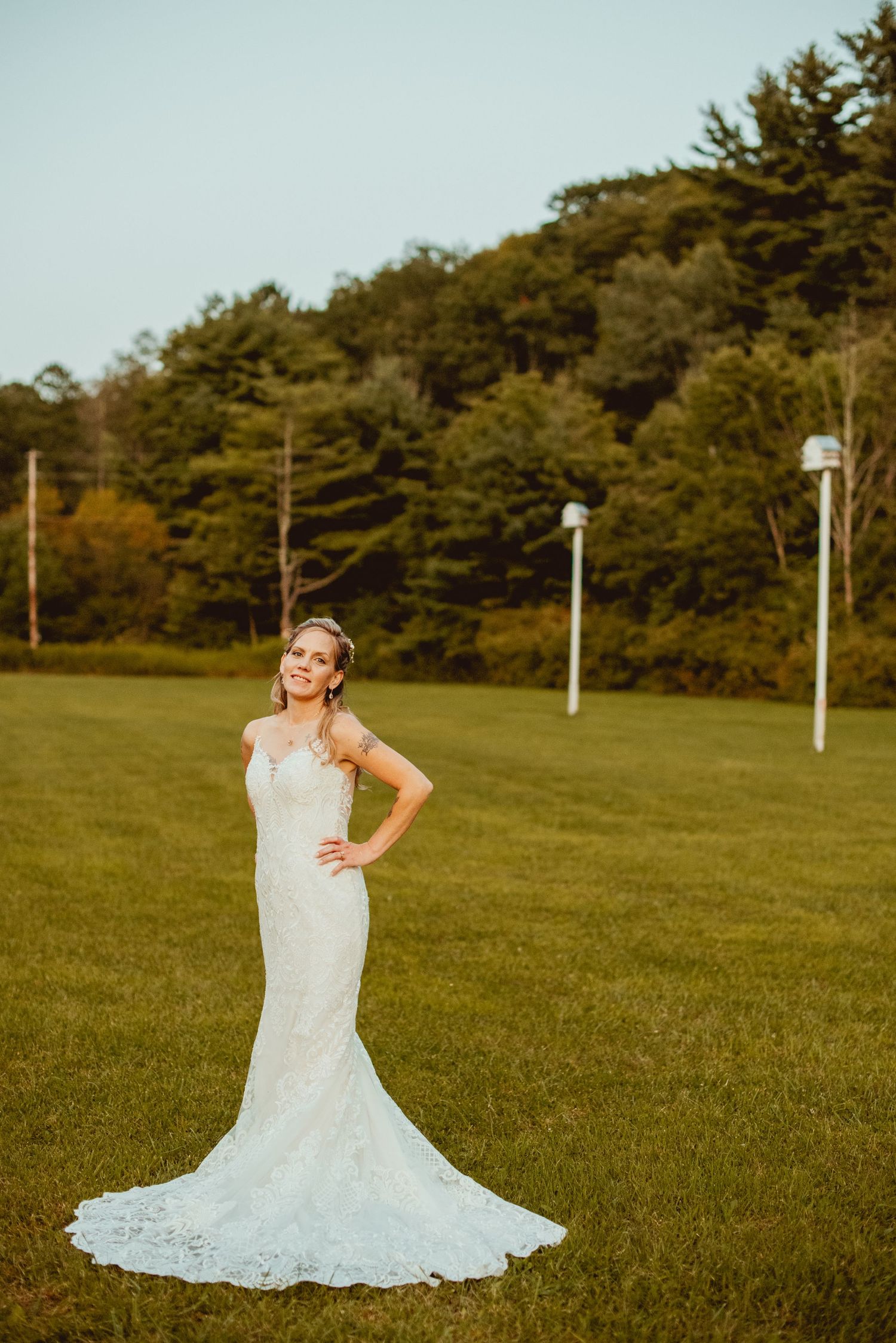 White lace wedding dress displayed on a grassy field with pine trees in the background.