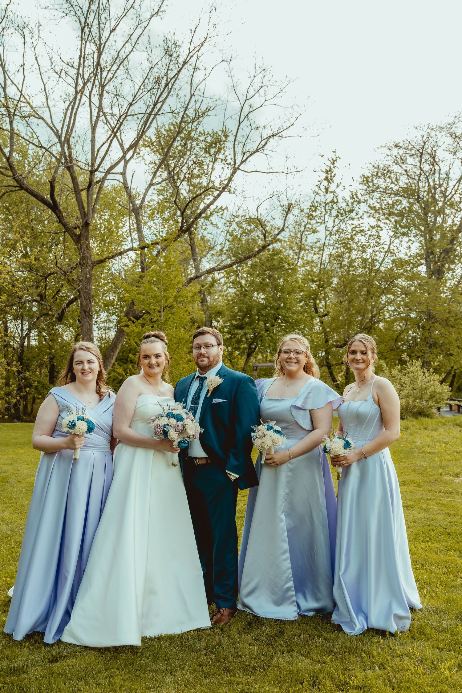 A groom in navy suit poses with four bridesmaids in light blue gowns holding bouquets on a green lawn.