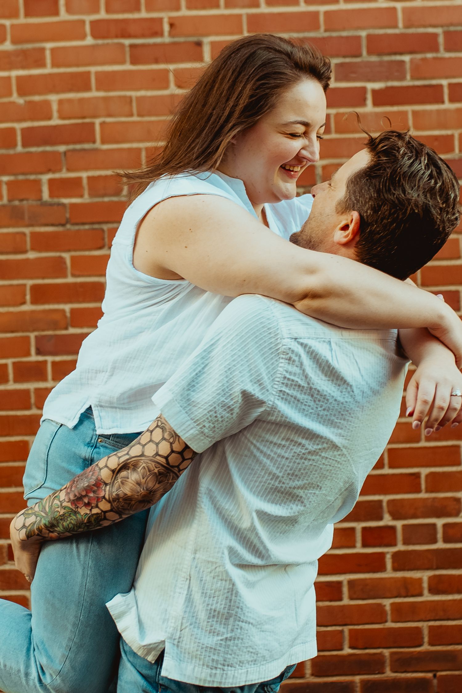 A joyful couple embraces against a brick wall, man with tattoos lifting smiling woman in casual outfits.