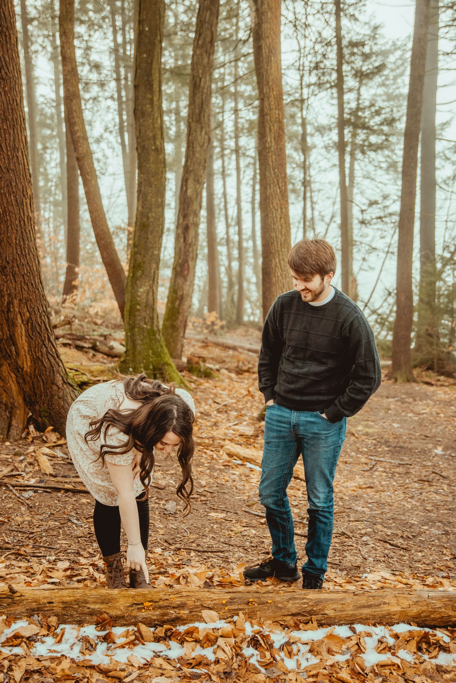 A couple stands together among tall pine trees on a forest floor covered in autumn leaves.
