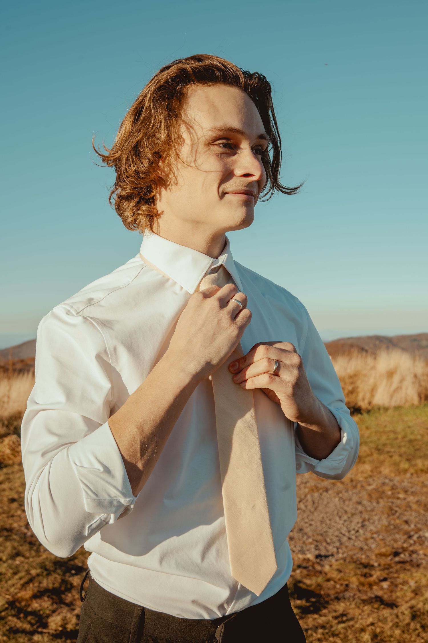 A person adjusts their tie against a desert landscape backdrop during golden hour.