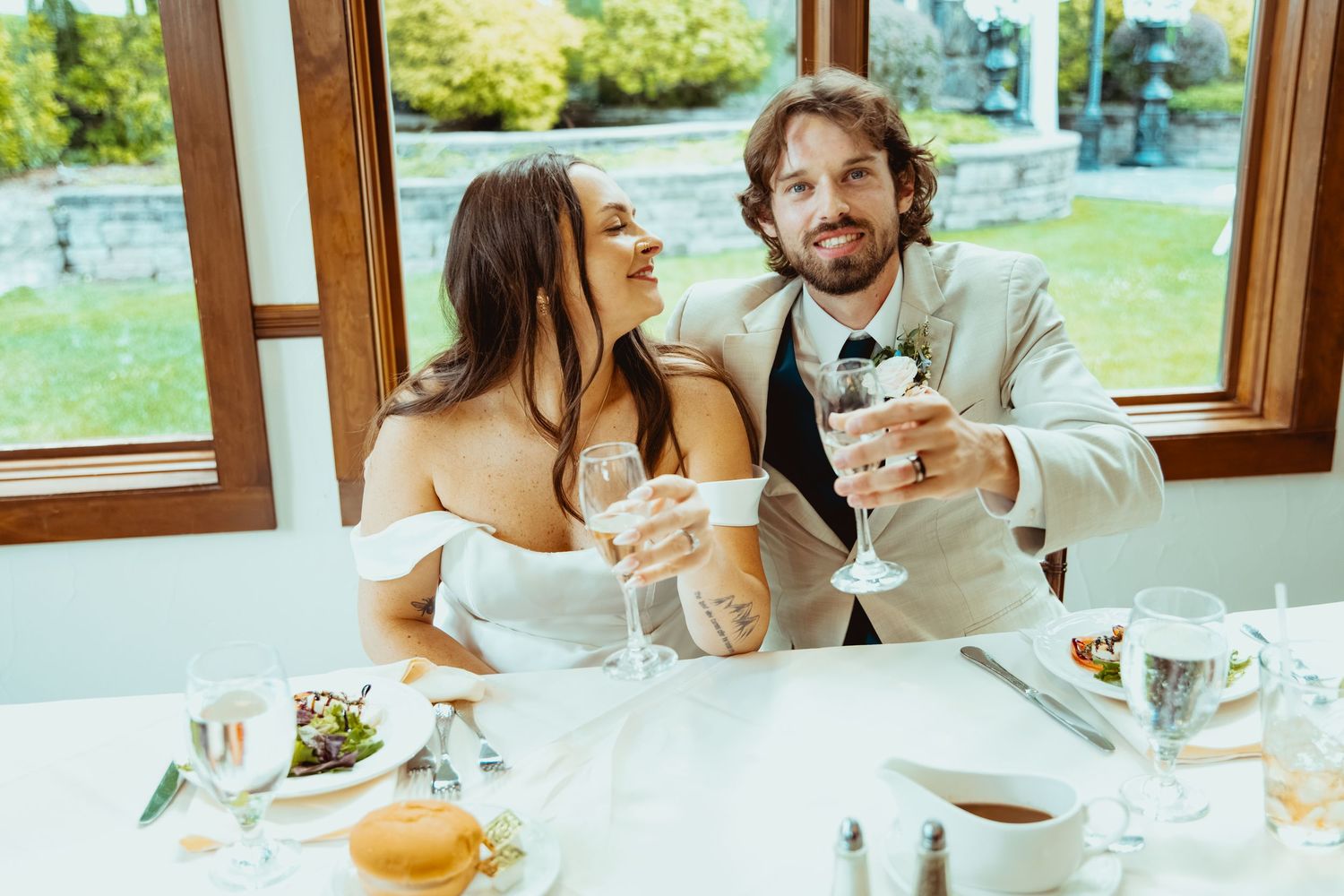 Smiling newlyweds in wedding attire toasting with wine glasses at reception dinner table.