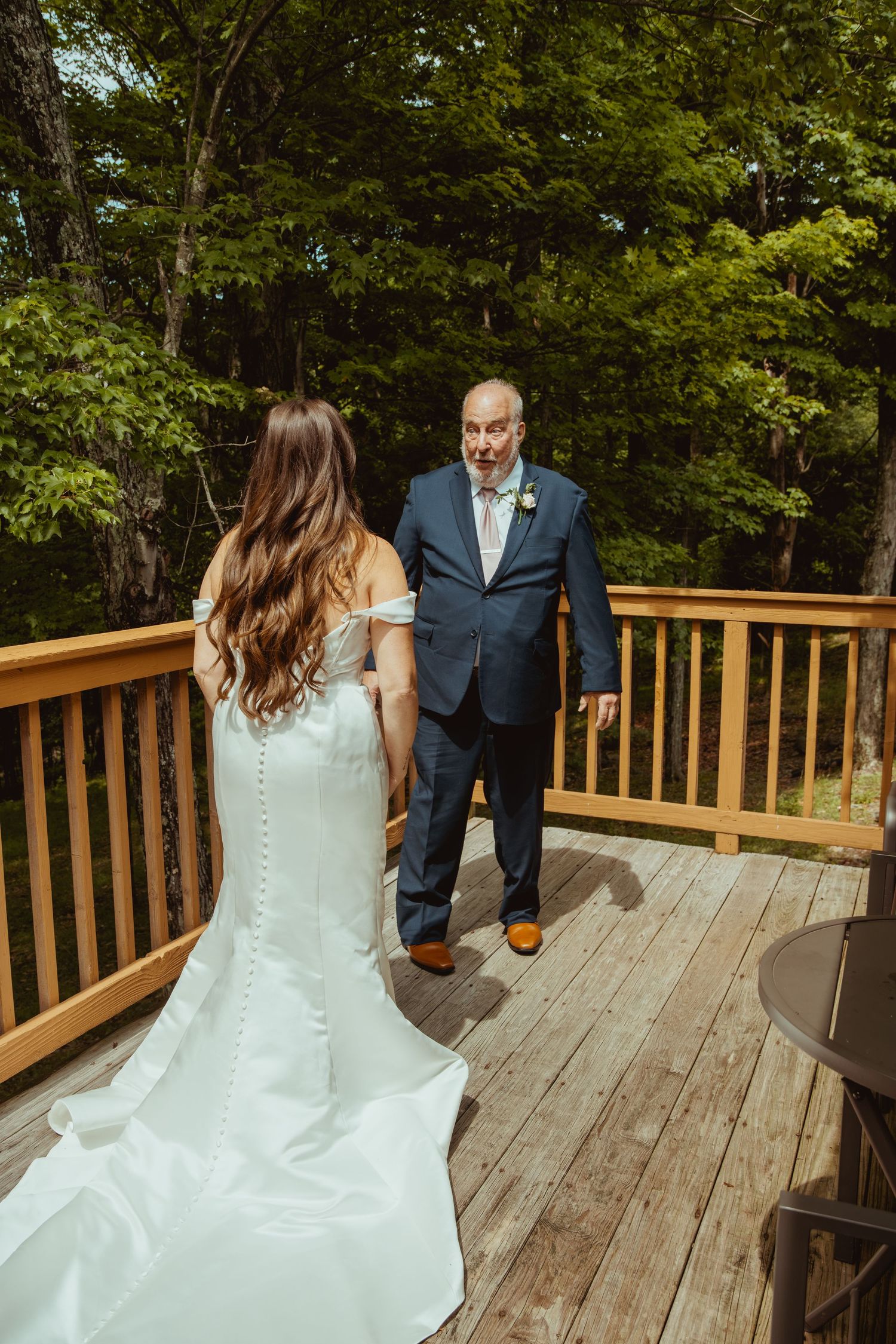 Bride in white gown shares first look moment with father on wooden deck surrounded by lush green trees.