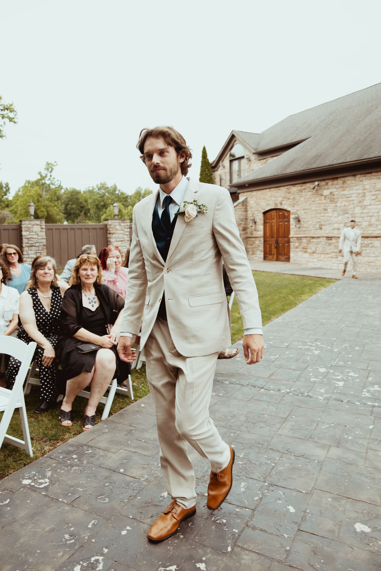 Groom walks down outdoor aisle in beige suit with boutonniere at garden wedding ceremony.