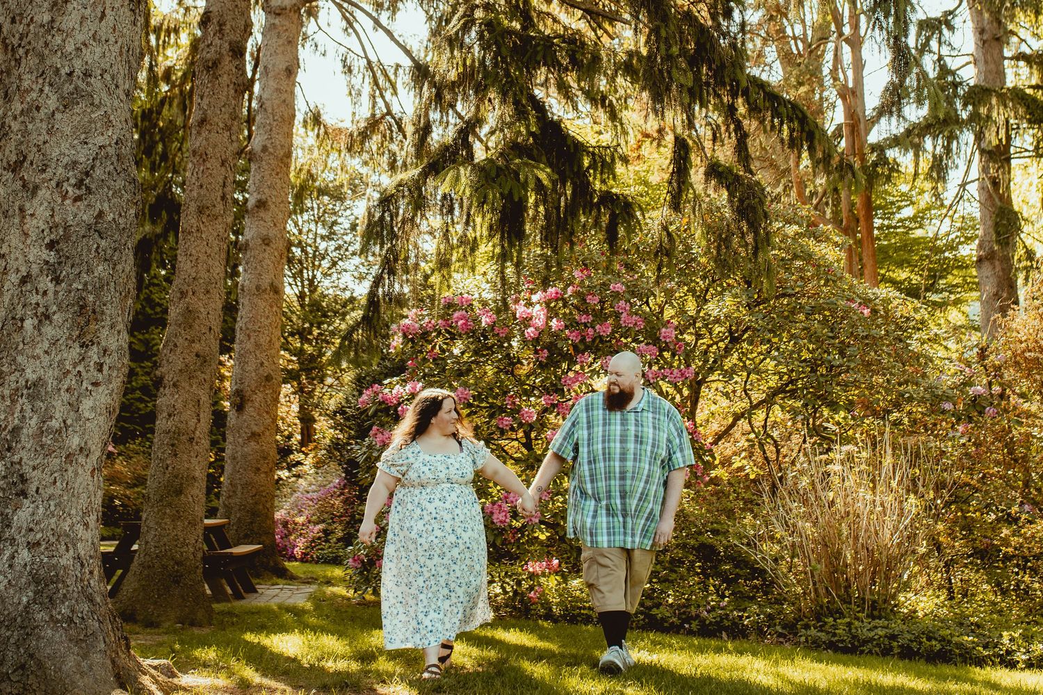 Couple holding hands walking through a lush garden with blooming pink flowers and tall trees.