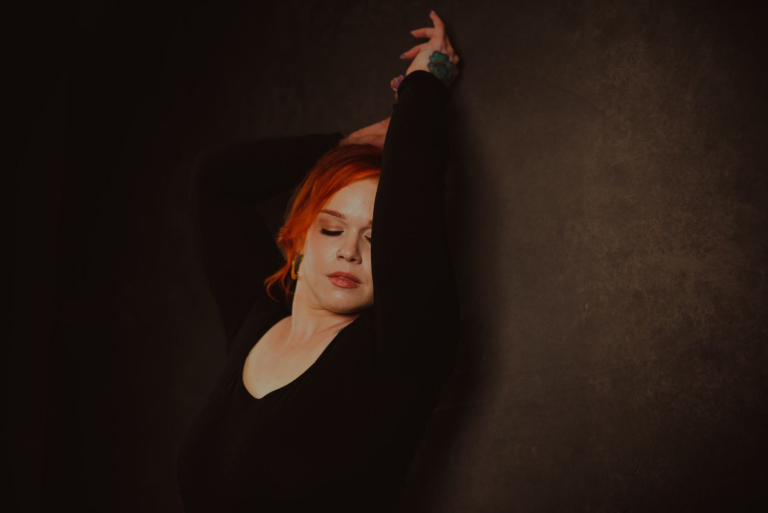 Red-haired woman in black top poses gracefully against dark wall with arm raised, eyes closed.