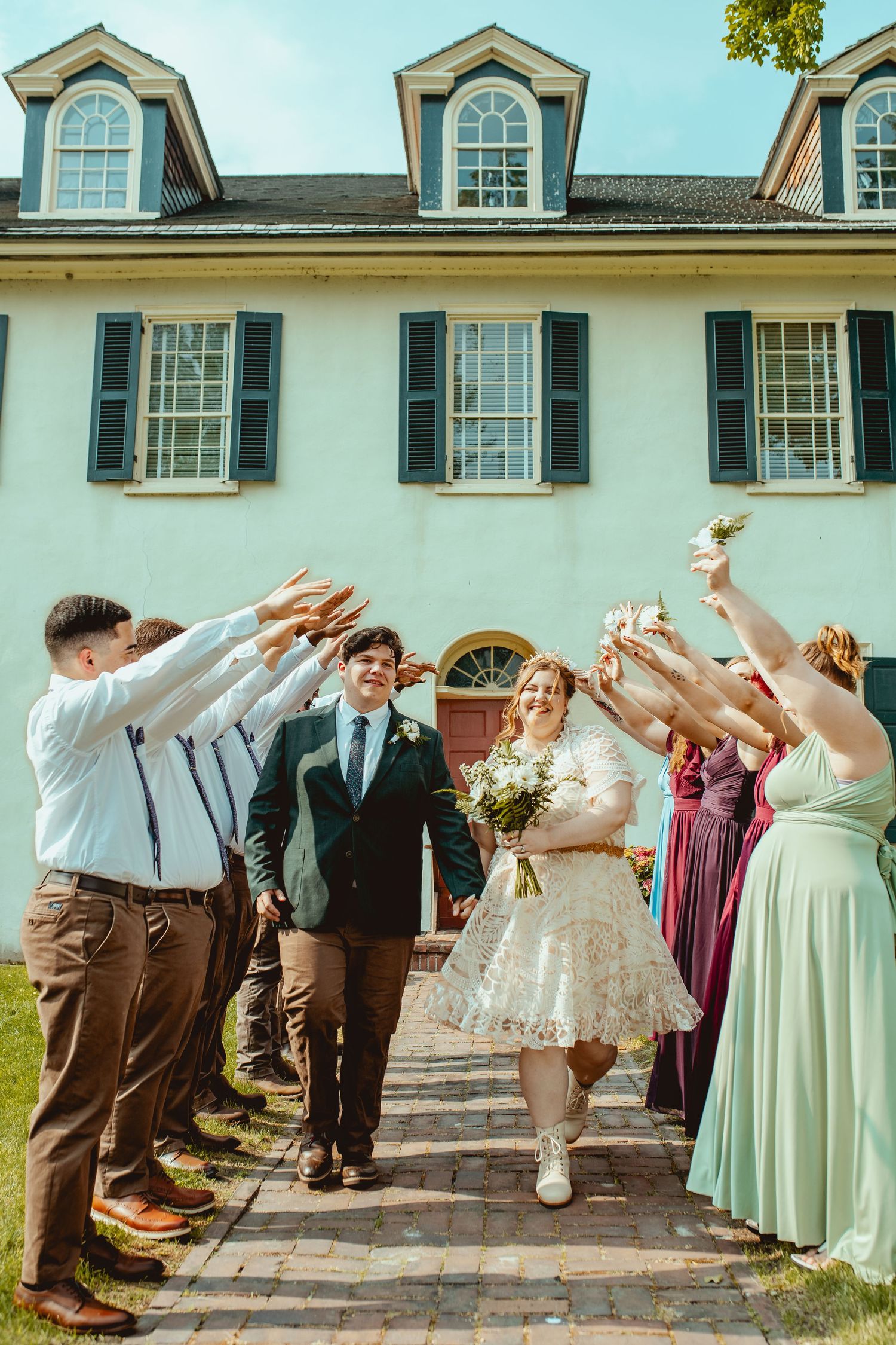 Wedding party members perform synchronized dance moves in coordinated outfits on a brick patio outside a white mansion.