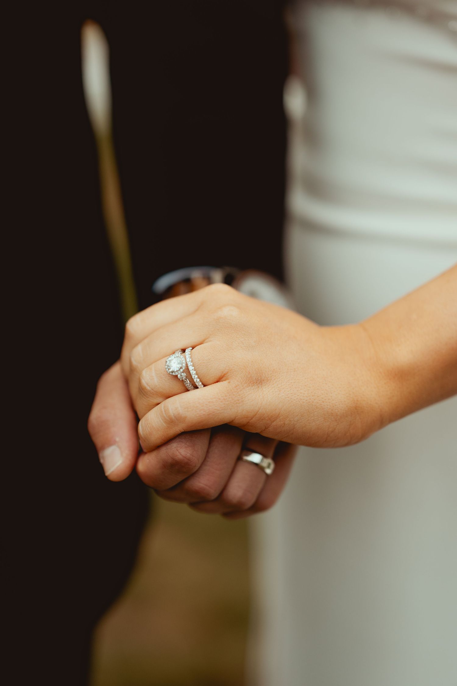 Close-up of bride and groom holding hands, showcasing a sparkling diamond engagement ring and wedding band.
