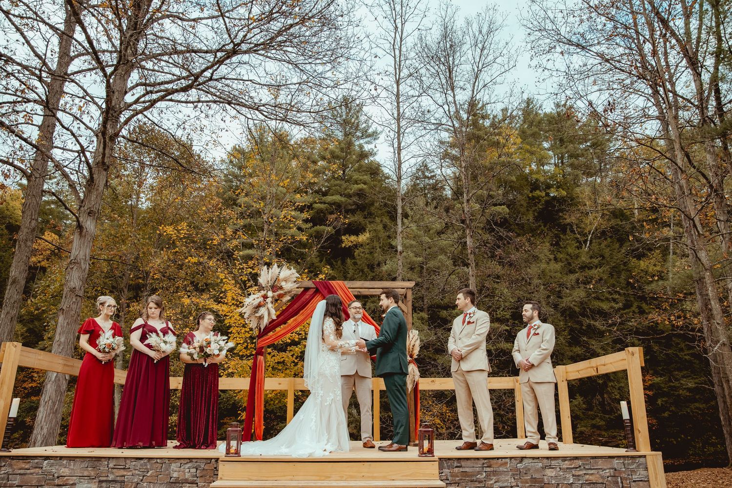 Outdoor fall wedding ceremony with bride, groom, and wedding party on wooden deck surrounded by autumn trees.