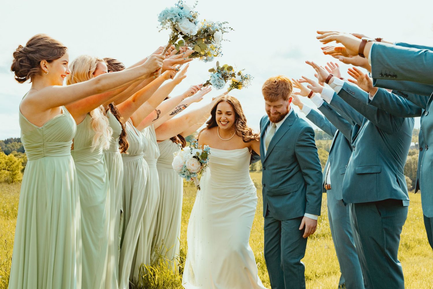 Celebration moment with wedding party in mint dresses reaching toward couple in sunny field.