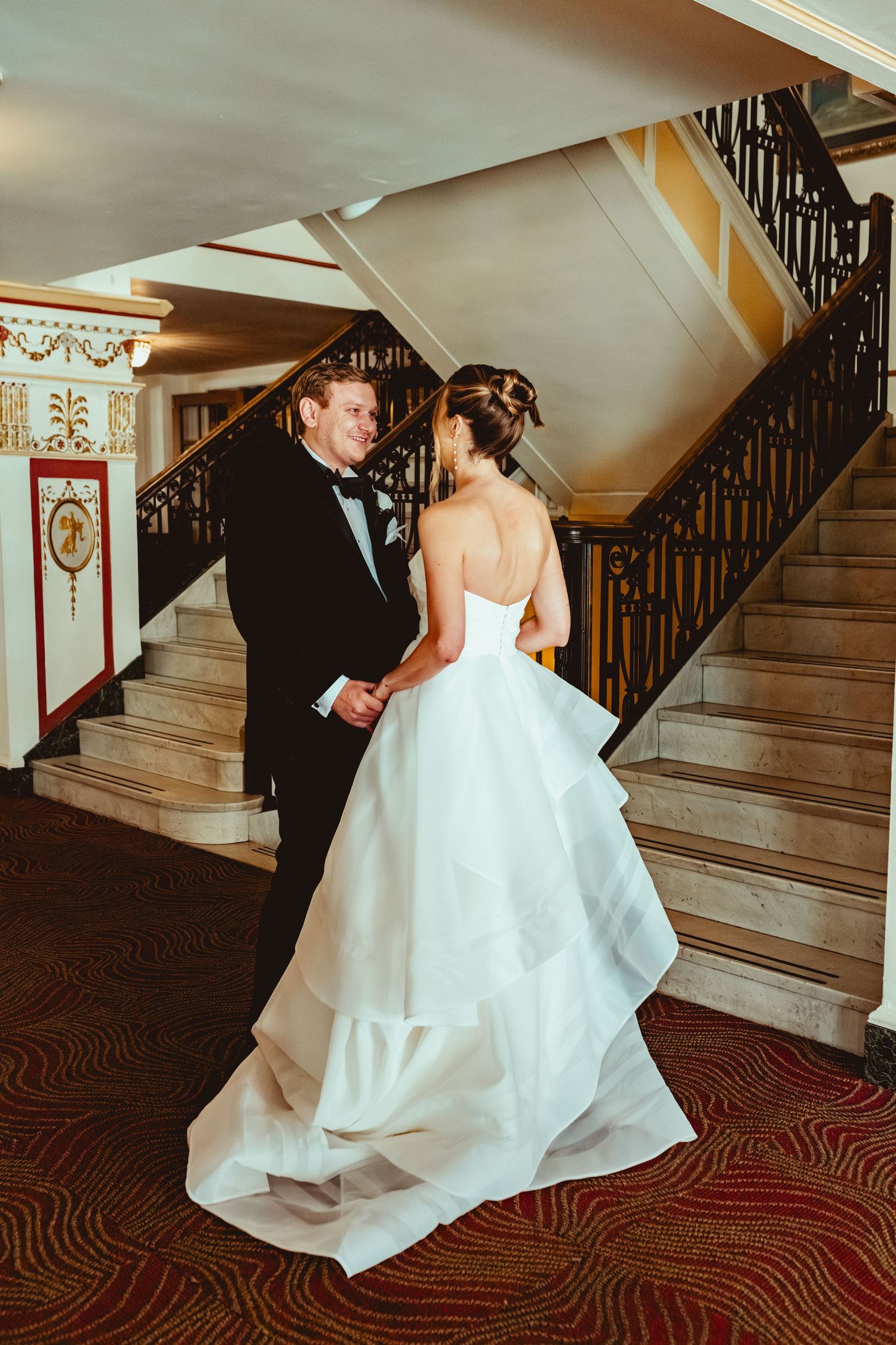 Bride in flowing white gown ascending grand staircase with ornate black railing.