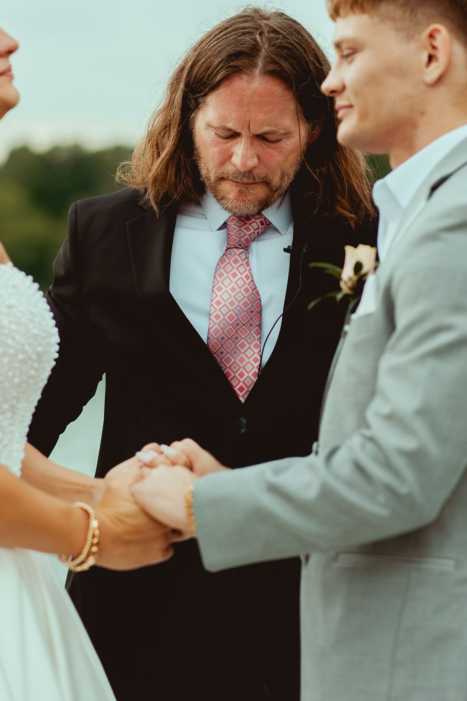 Wedding officiant with long hair holding hands with bride and groom during outdoor ceremony.