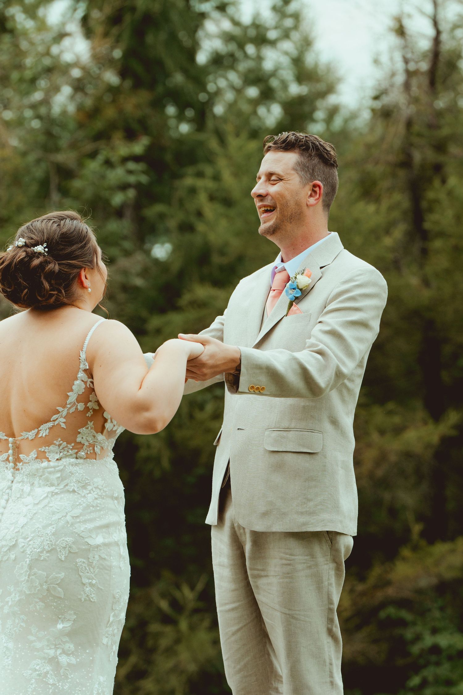 Smiling groom in tan suit holding bride's hands during outdoor first look wedding moment.