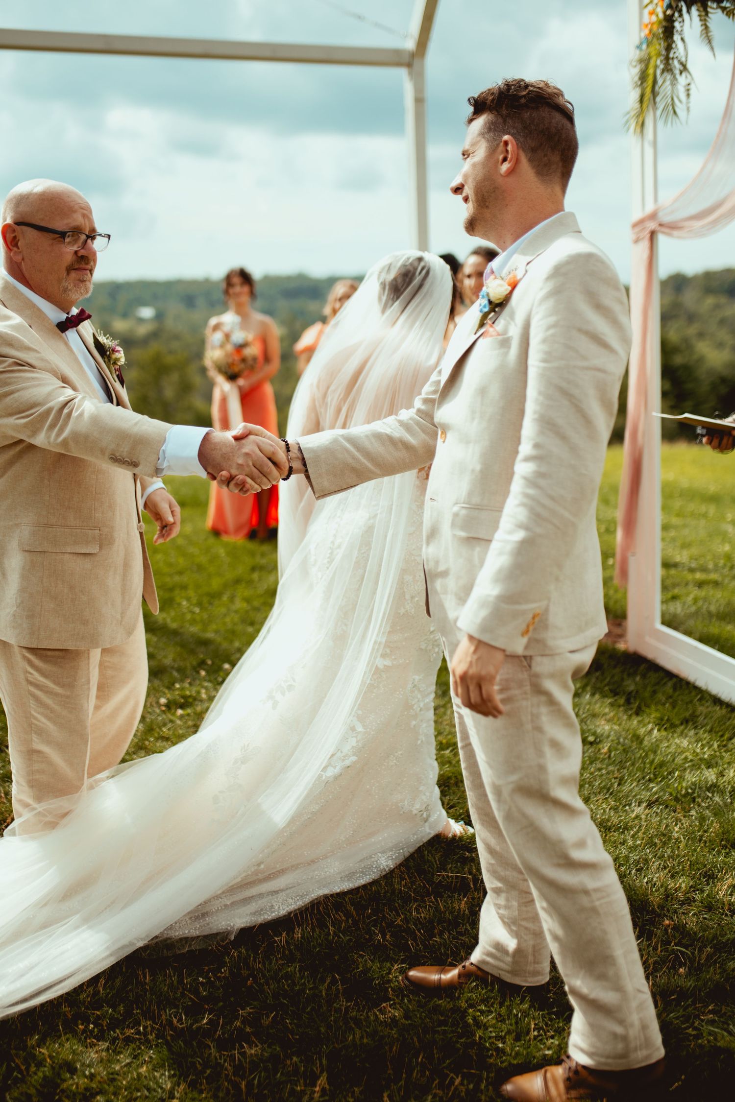Father of the bride shakes hands with the groom during an outdoor wedding ceremony on a lush green hillside.