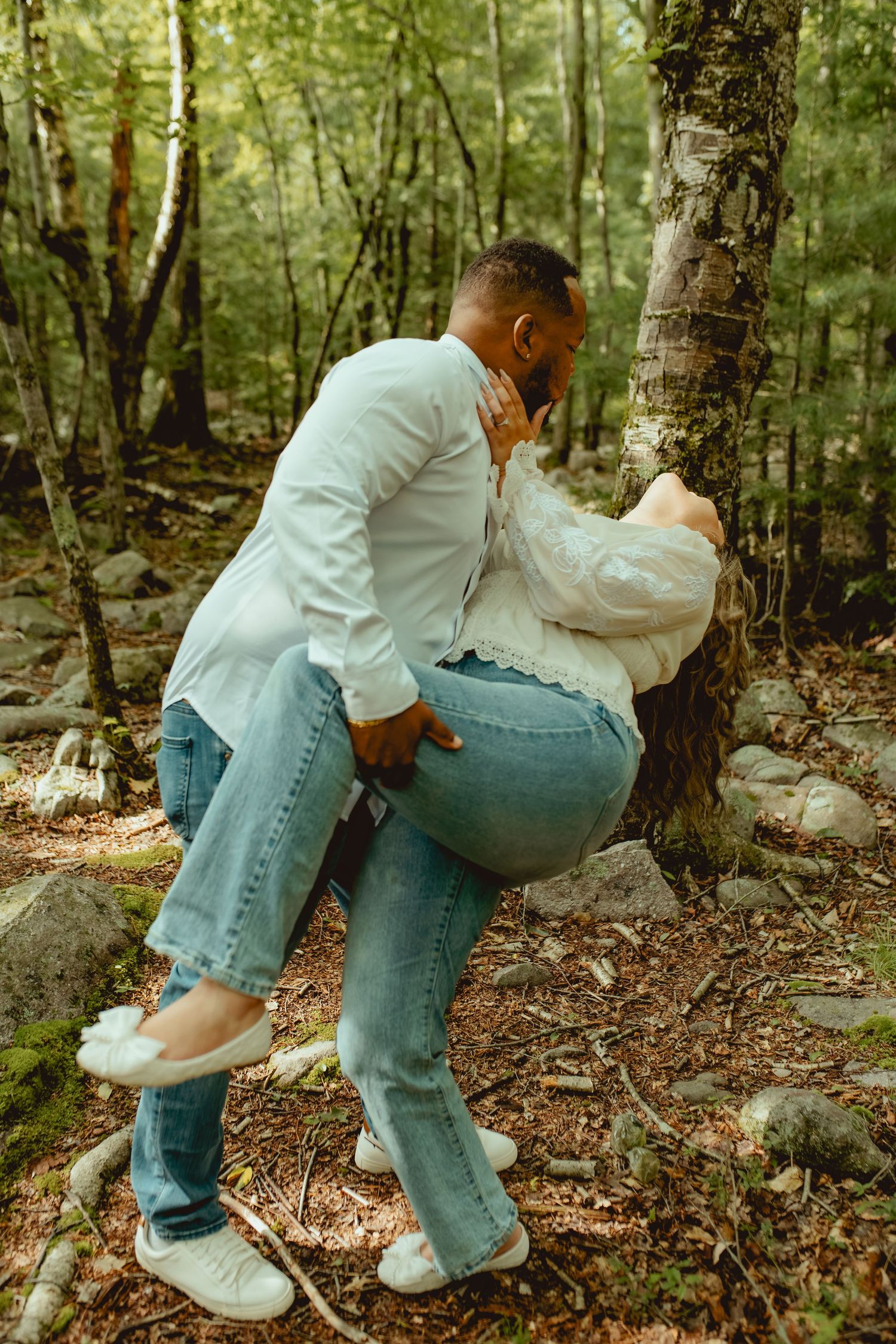 Couple sharing a romantic kiss against a birch tree in a lush green forest, both wearing white shirts and jeans.