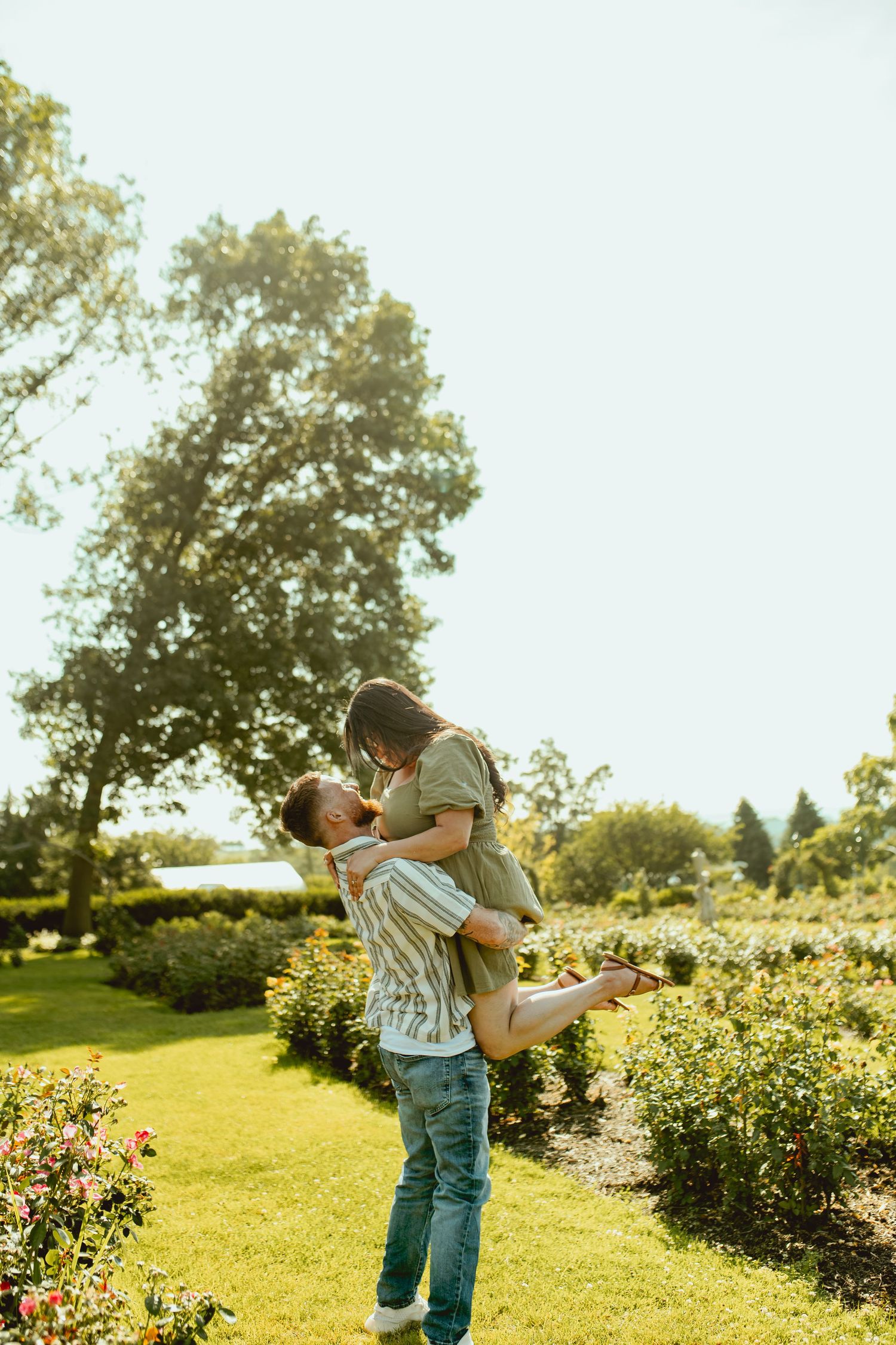 A couple sharing a joyful moment in a sunny garden, man lifting woman among colorful roses and lush green trees.