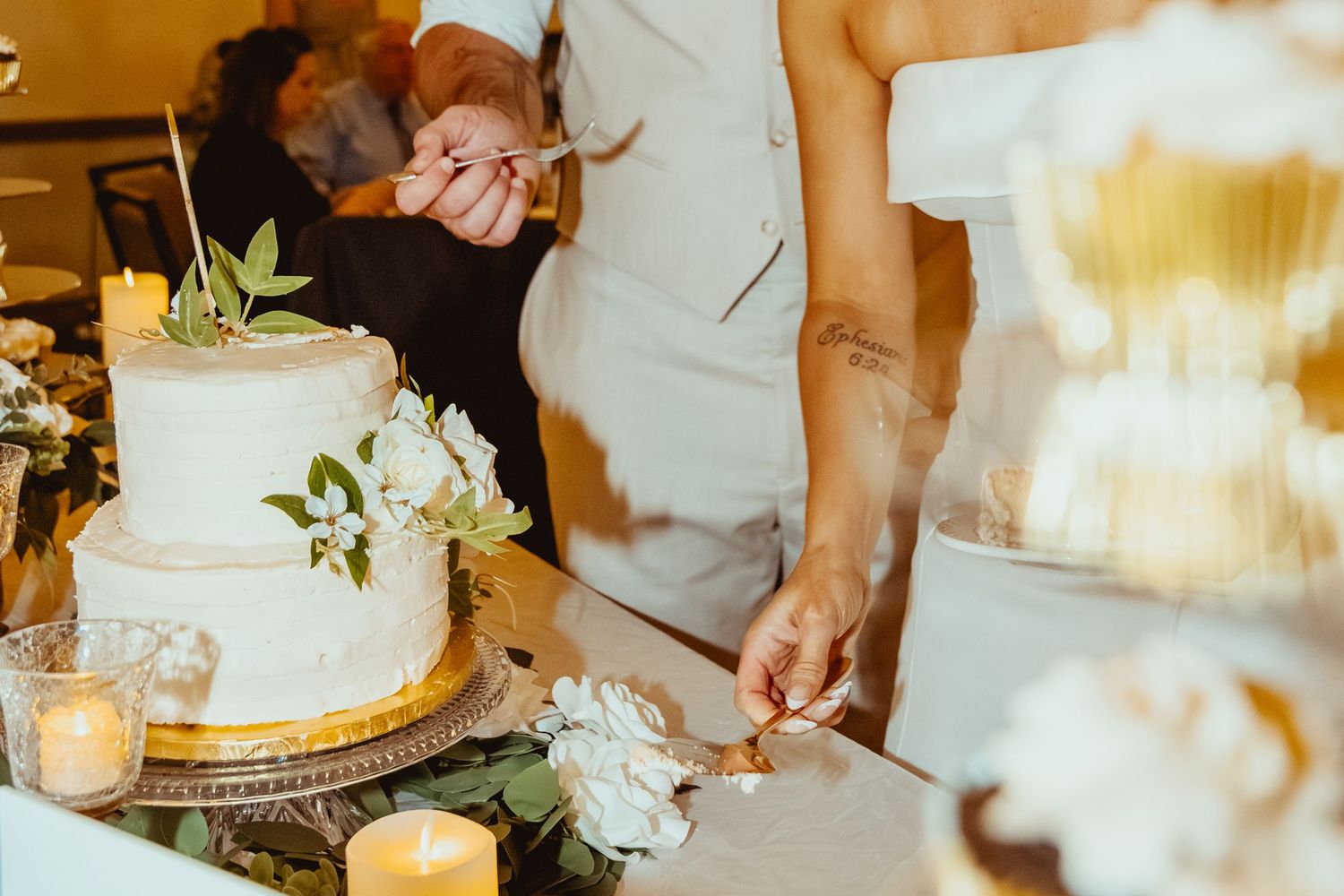 Close-up view of hands cutting a white wedding cake with gold accents and floral decorations.
