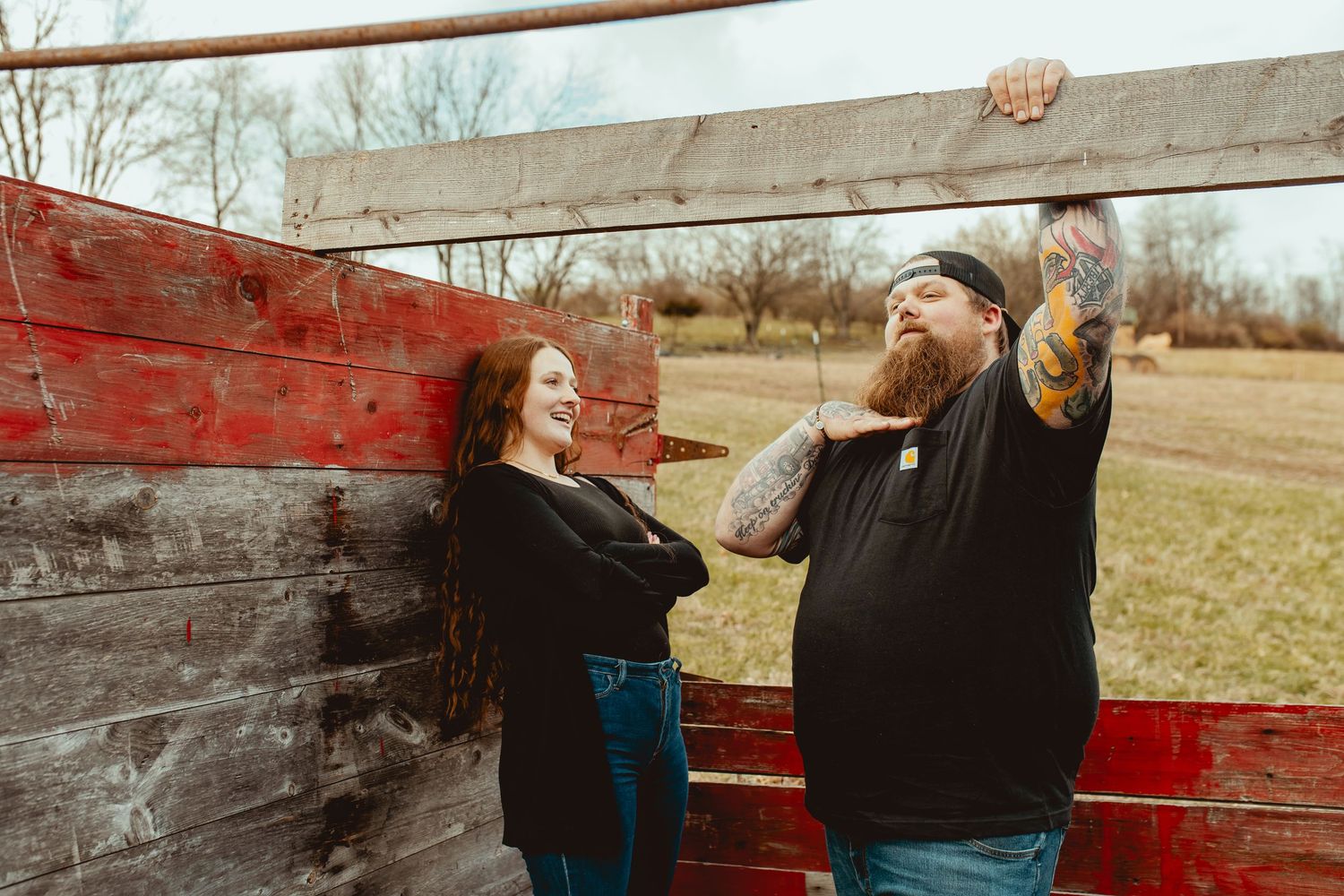 A bearded tattooed man and a woman laugh together leaning against a rustic red wooden fence outdoors.