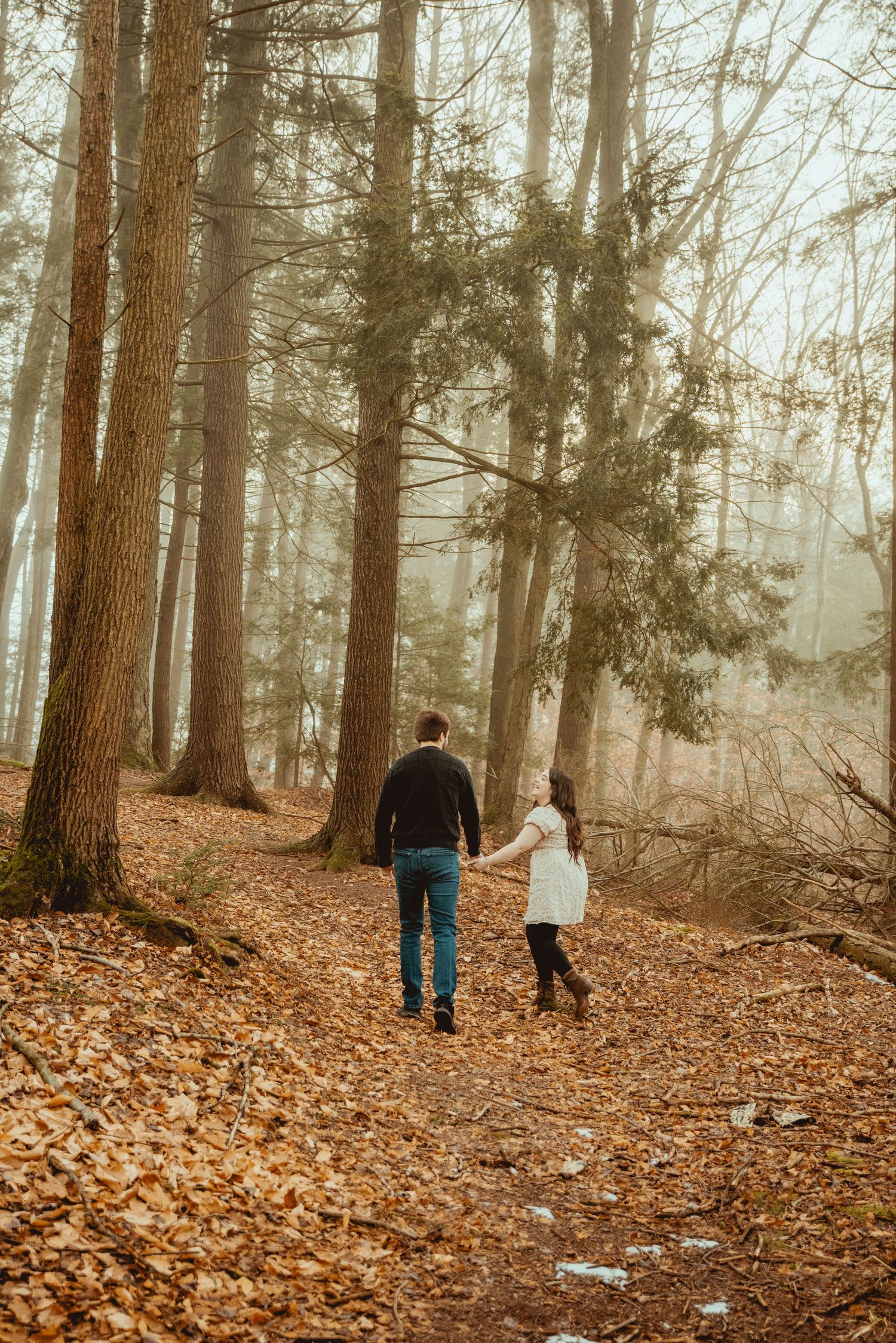 People walk through a foggy forest trail covered in fallen autumn leaves with tall tree trunks surrounding them.