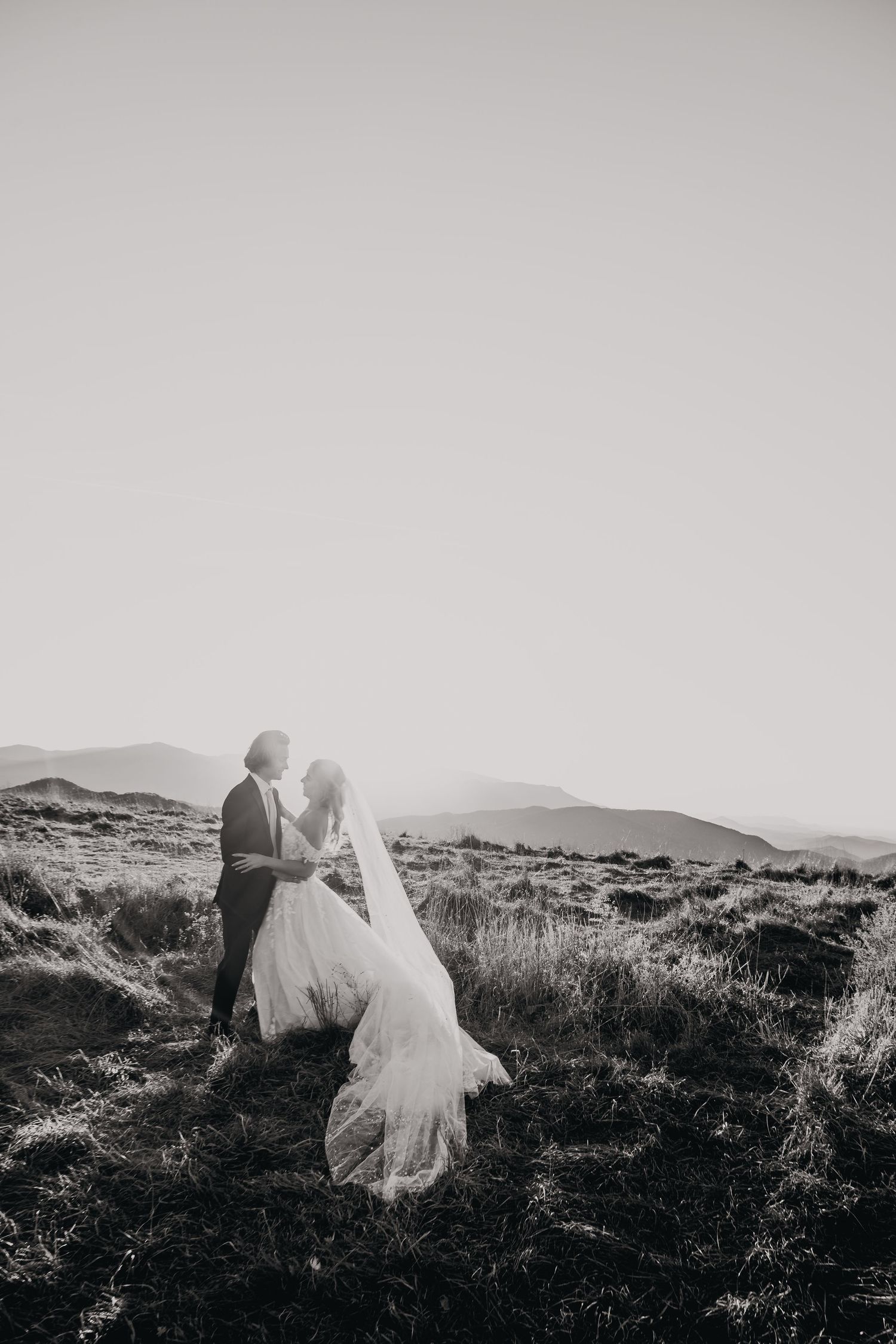 A black and white wedding portrait of a couple embracing on a hilltop landscape.