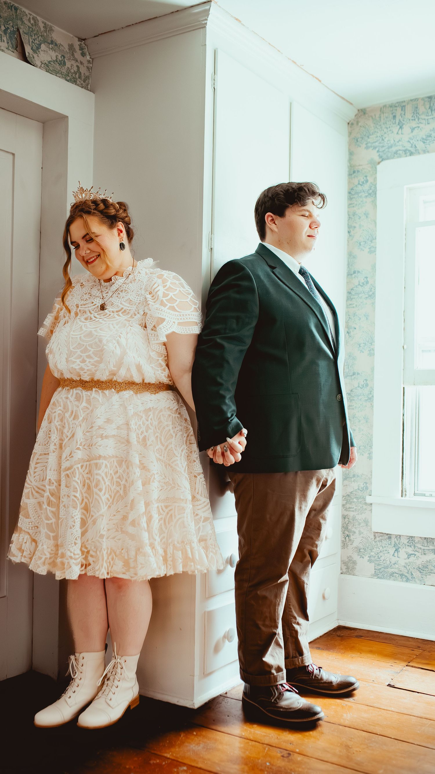 A romantic first look moment between a bride in a short lace dress and groom in a dark suit near a window.