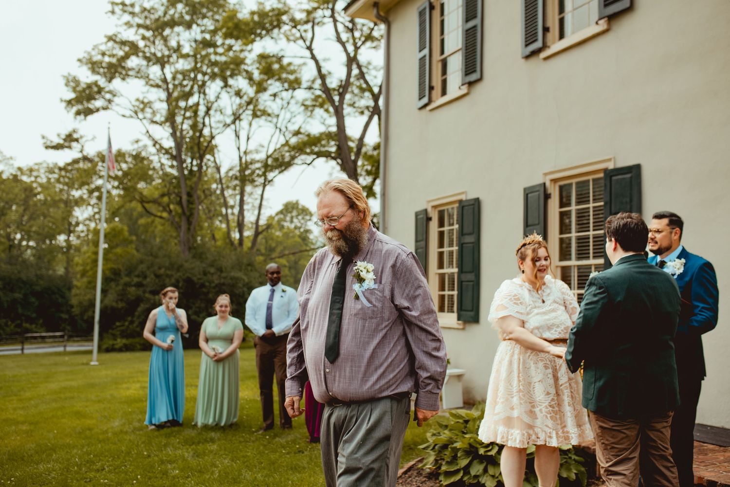 Wedding party members gather on the lawn outside a white building during an outdoor celebration.