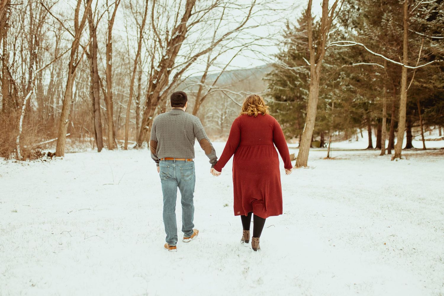 A couple walks hand in hand through a snowy winter landscape in a park with bare trees.