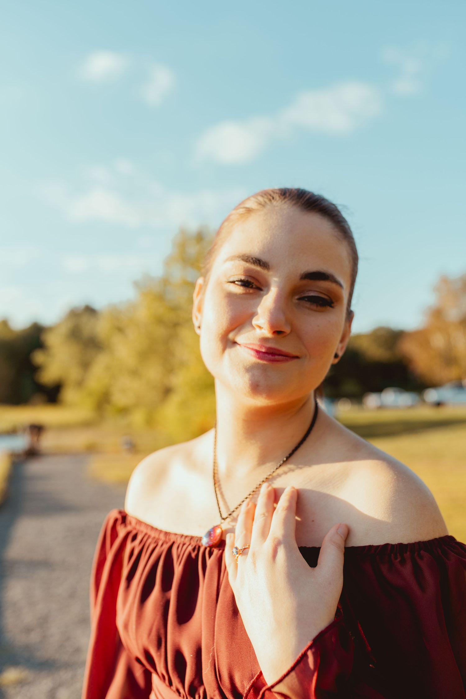 A person in a burgundy top poses outdoors on a sunny autumn day with trees in the background.