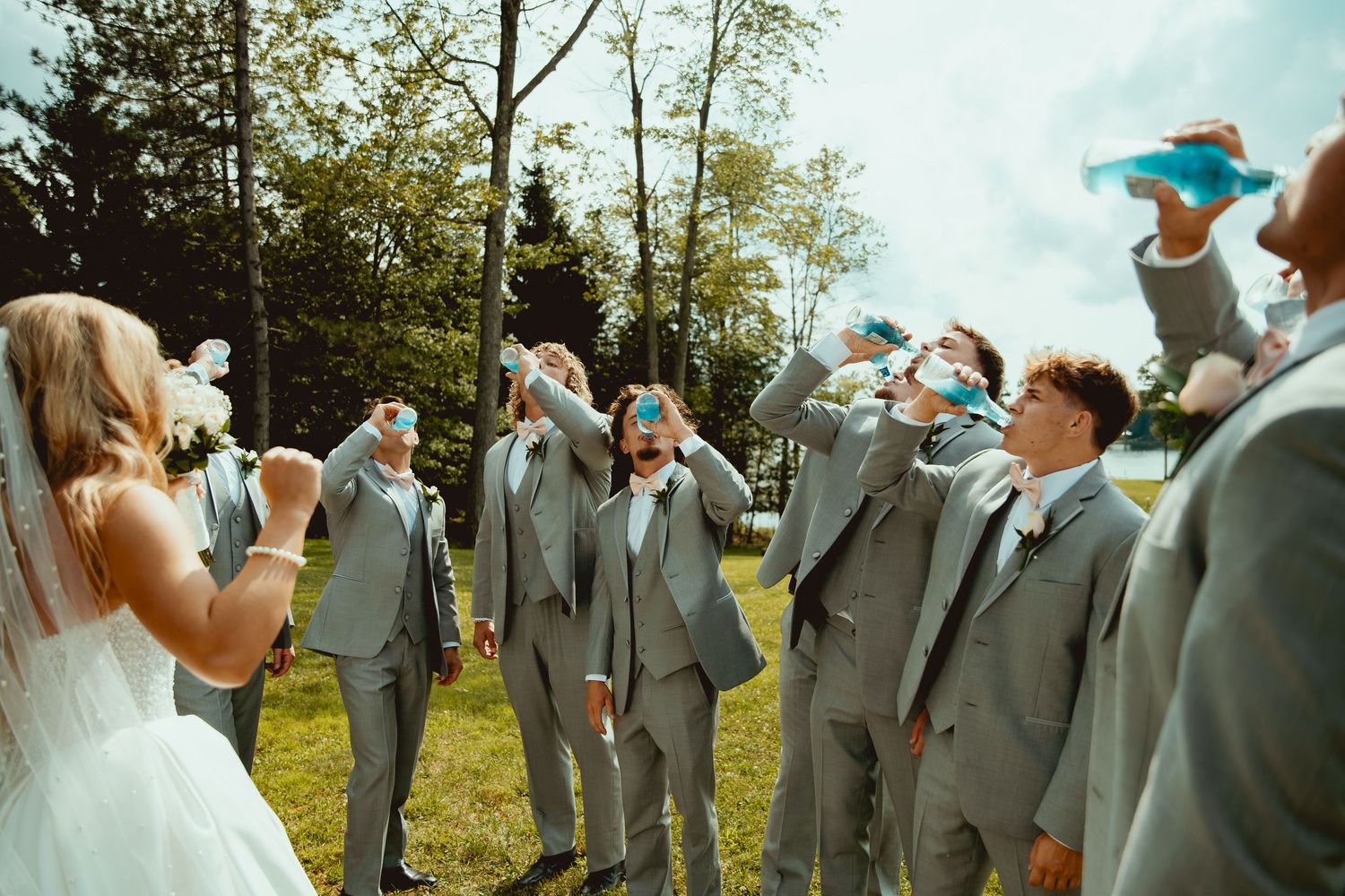 Wedding party in gray suits toasting with blue drinks outdoors, bride in white dress celebrating with groomsmen on sunny day.