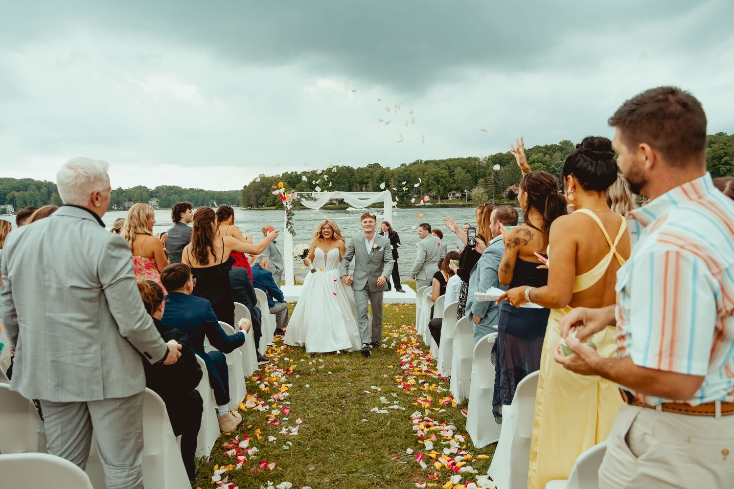 Newlyweds walk down outdoor aisle as guests toss petals by a scenic lakeside wedding ceremony.