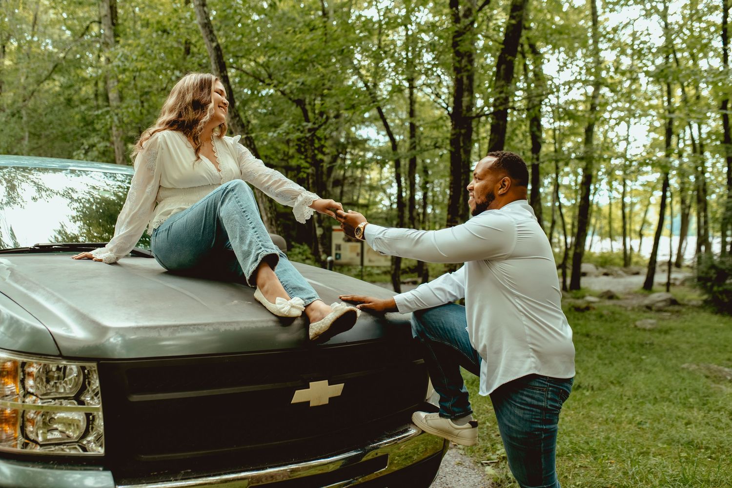 Couple sharing a romantic moment on the hood of a black Chevrolet truck in a lush green forest setting.