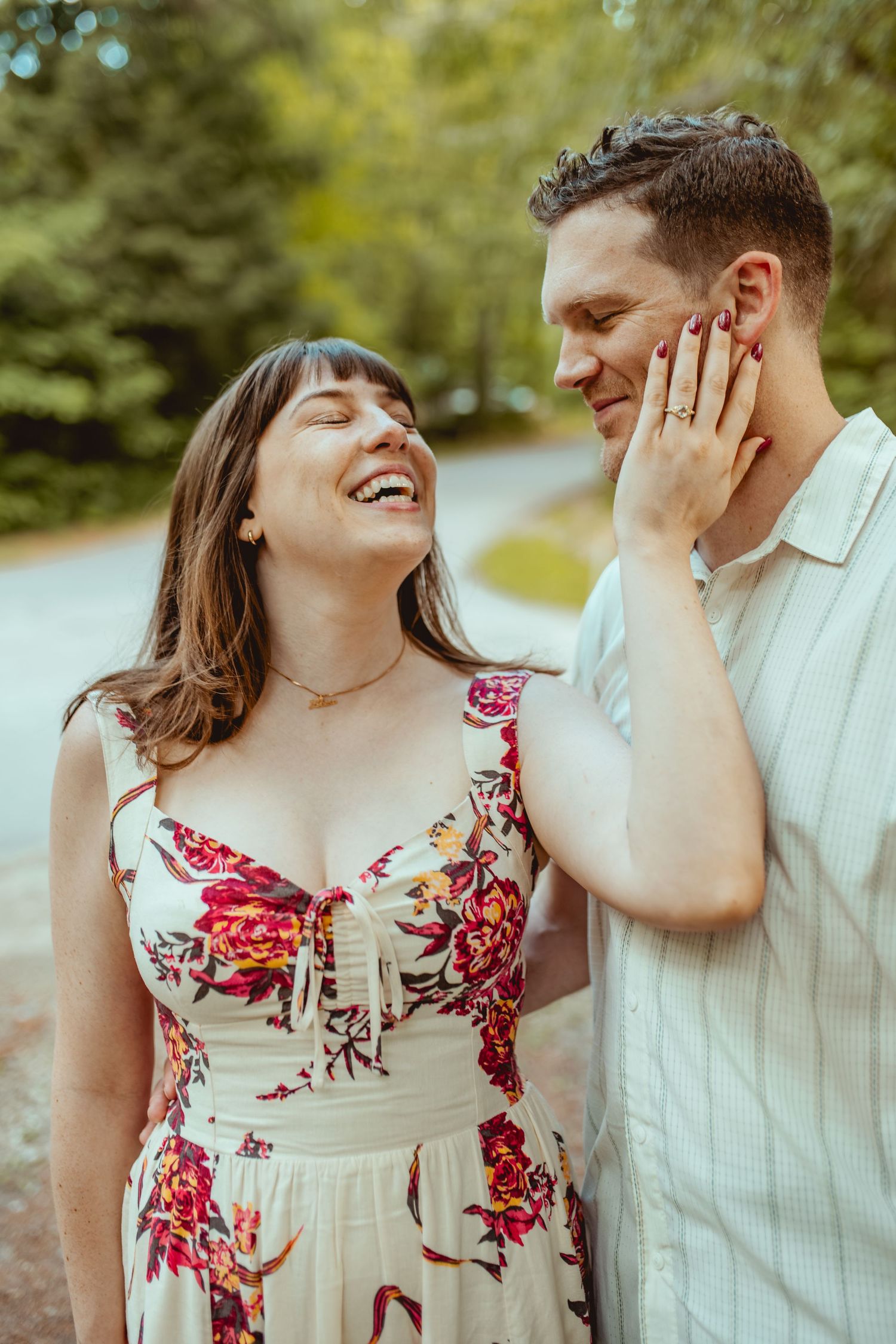 A laughing woman in a floral dress tenderly touches a smiling mans face outdoors on a tree-lined path.