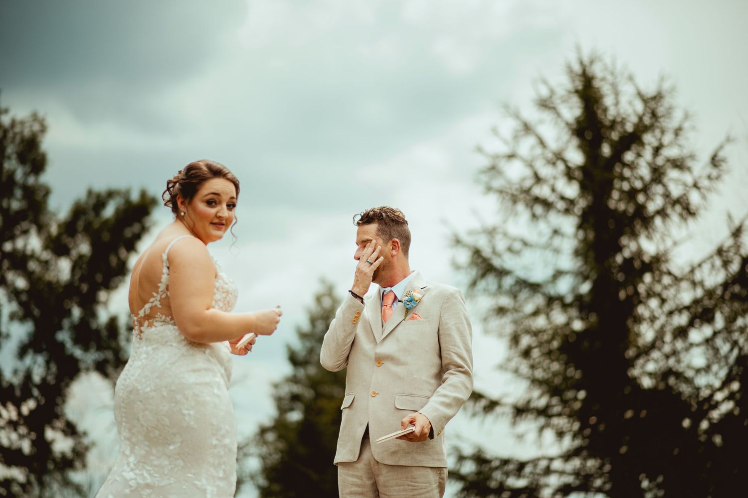 Emotional groom wipes tears as bride smiles during outdoor wedding ceremony with trees behind.