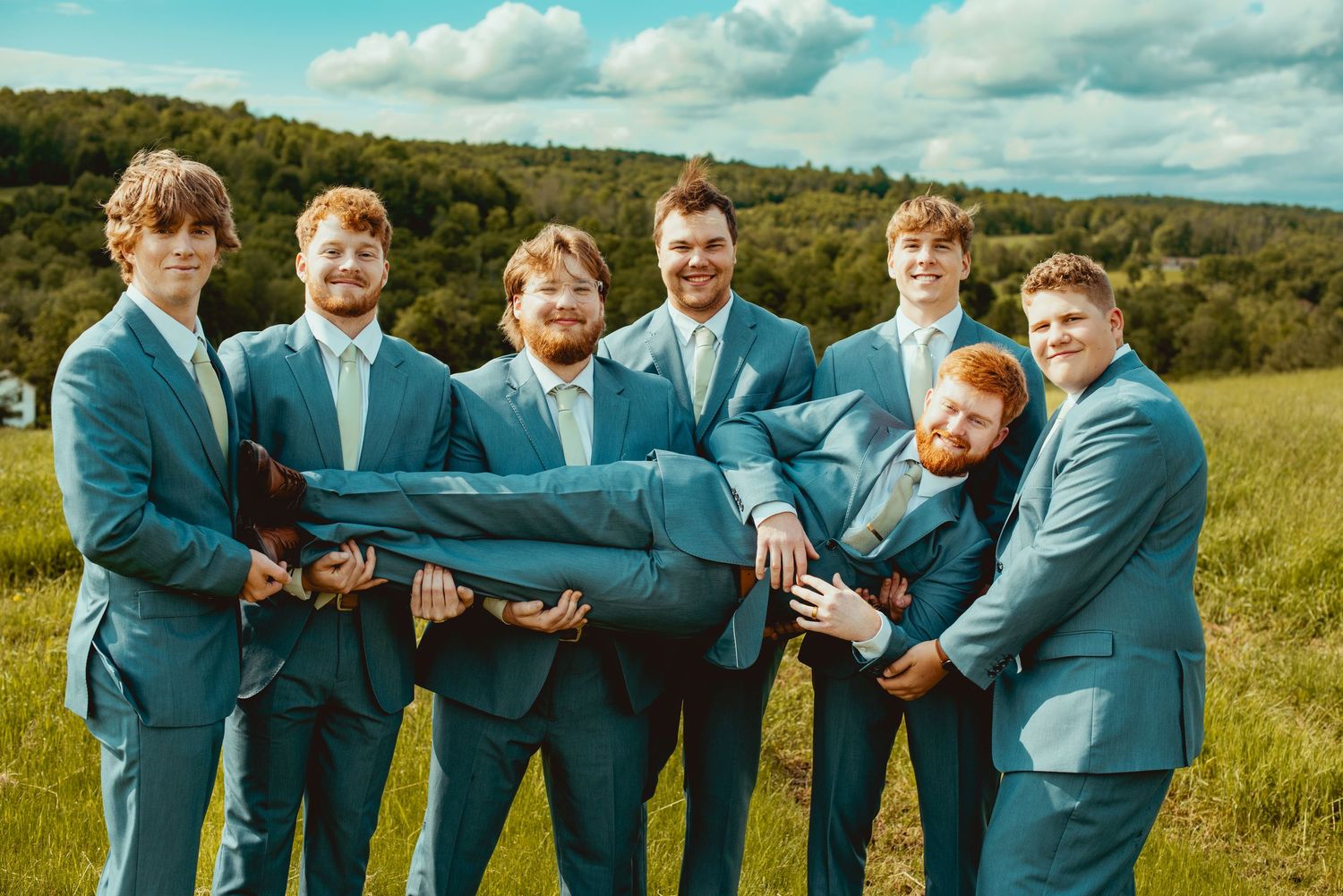 Groomsmen in teal suits playfully carrying one of their own in outdoor field.