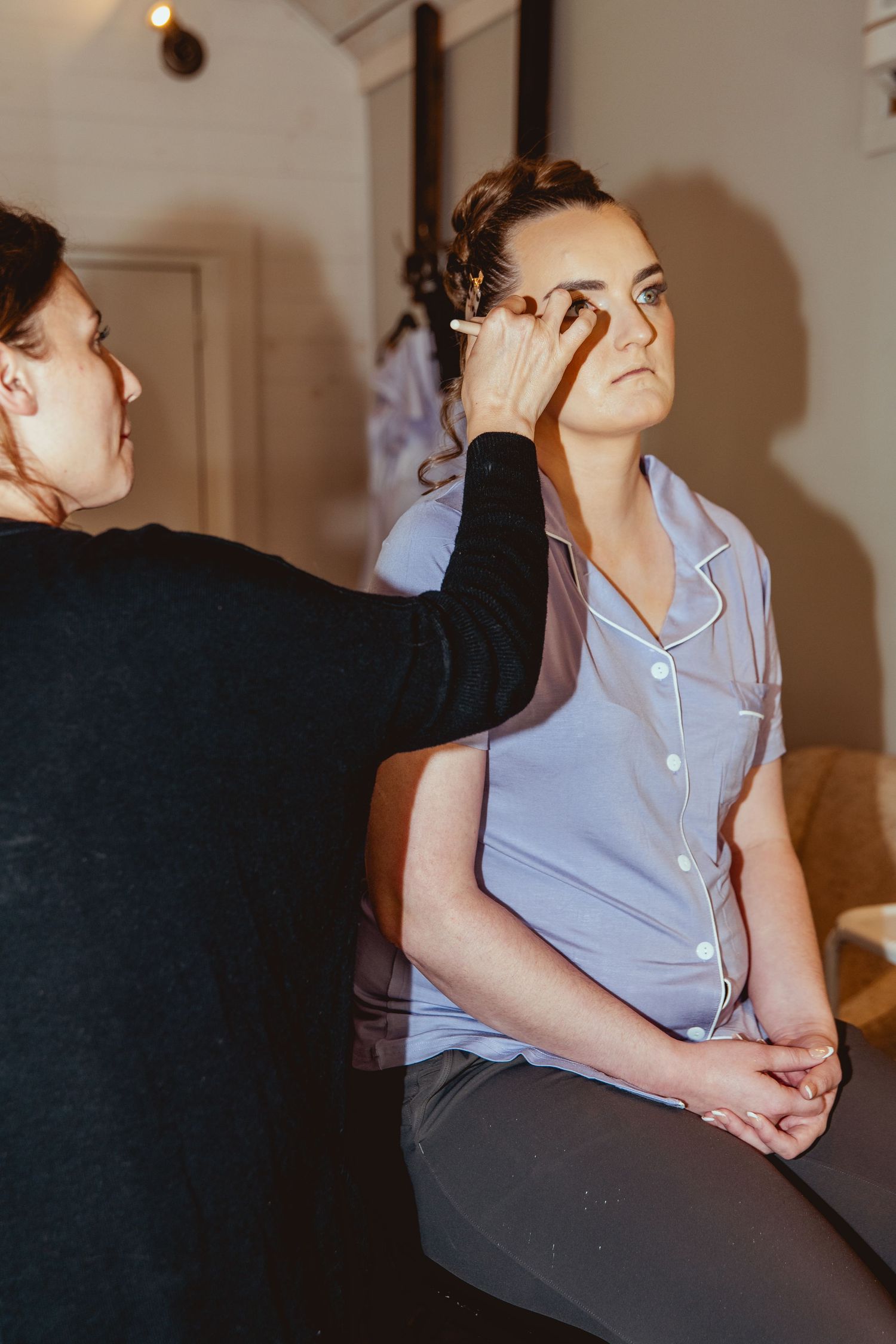 Professional makeup artist applying eye makeup to bride wearing light blue satin pajamas before wedding ceremony.