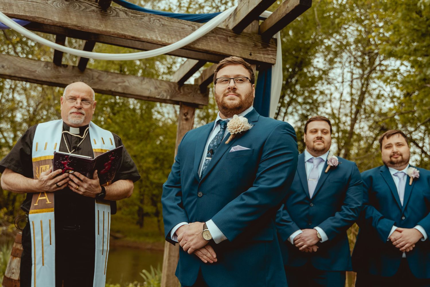 Groom in navy suit stands at outdoor wedding altar with groomsmen and officiant holding Bible among trees.