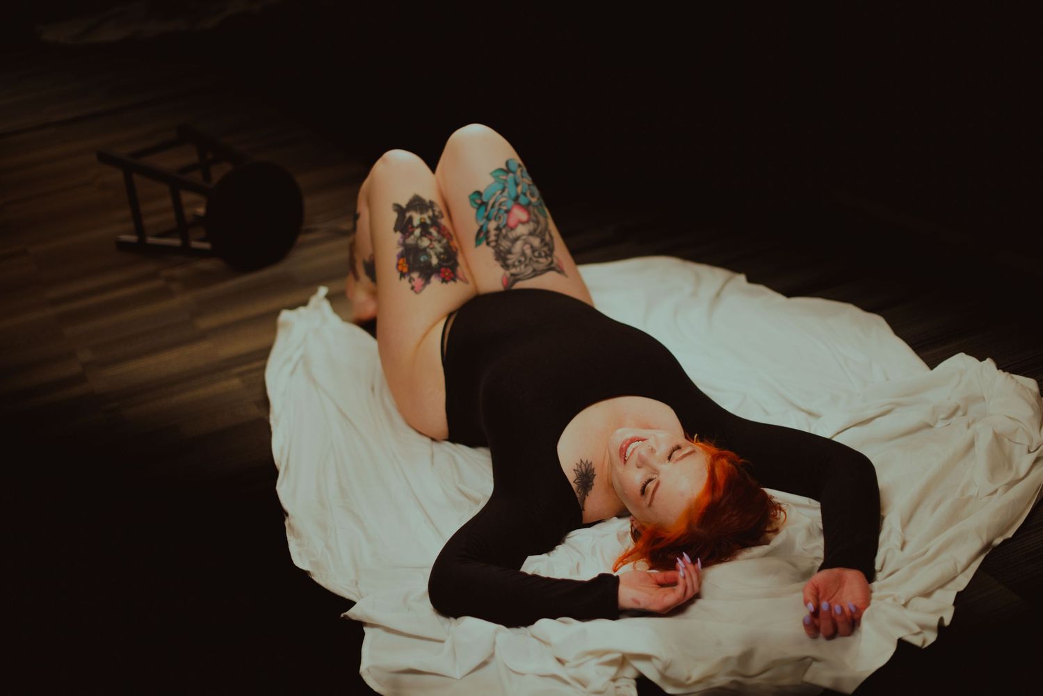 Tattooed redhead woman lying on white fabric on dark floor in black bodysuit, colorful floral tattoos on legs.