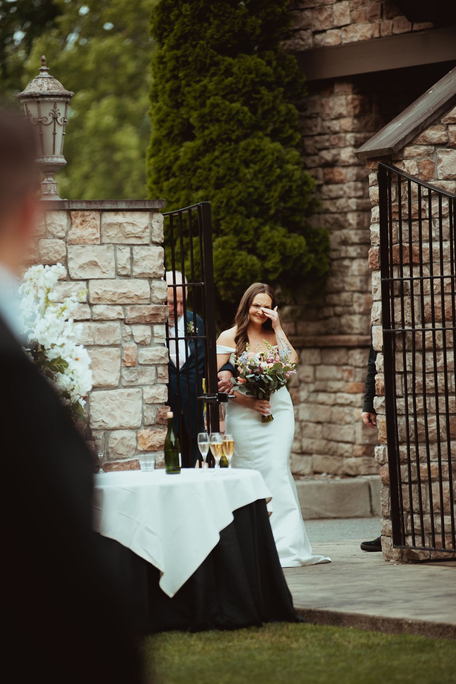 Emotional bride in white off-shoulder gown holds bouquet while walking through stone venue entrance.