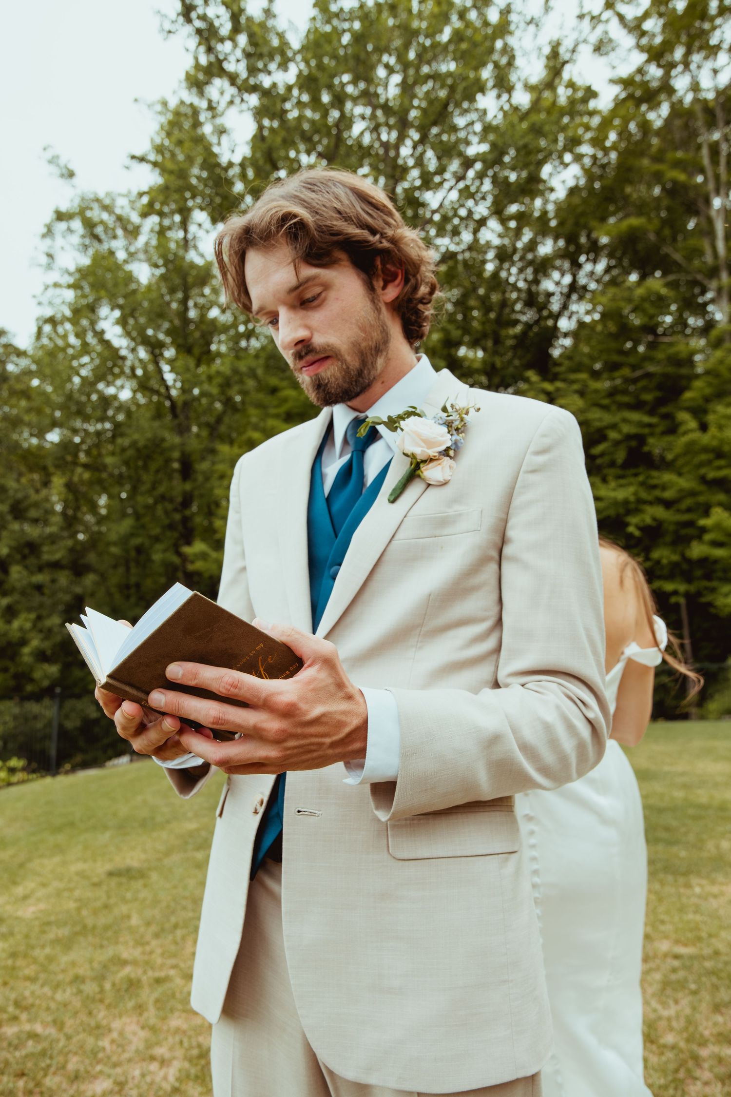 Groom in beige suit with blue tie reads vows from leather book outdoors at wedding ceremony.