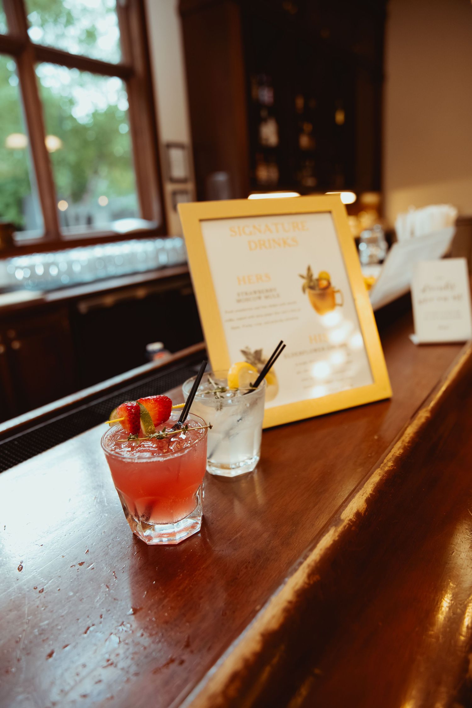 Two signature cocktails displayed on wooden bar beside gold-framed drink menu at wedding reception.