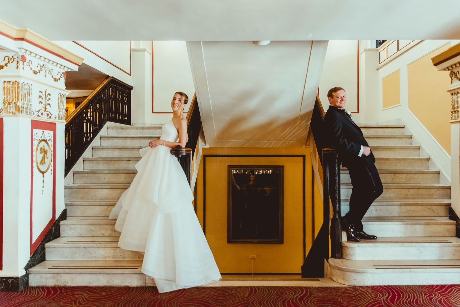 Wedding couple poses dramatically on grand marble staircase with classic architectural details.