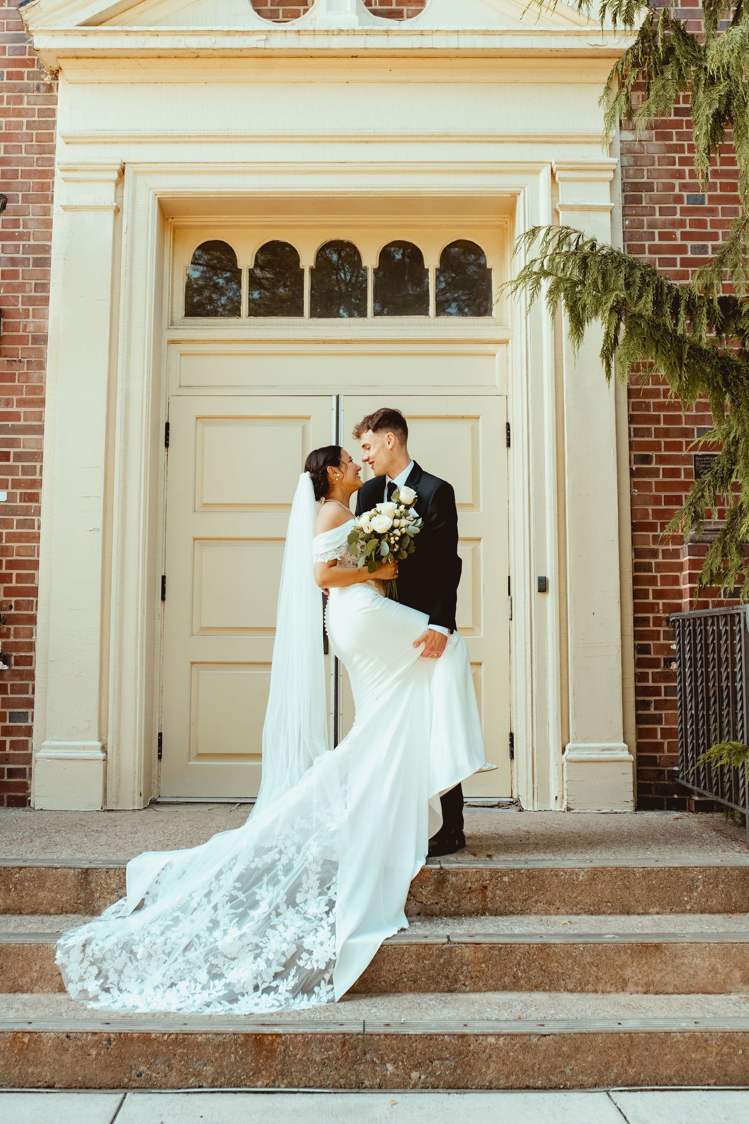 Bride in a lace-trimmed gown and groom in a black suit embrace on brick church steps in front of tall cream double doors.