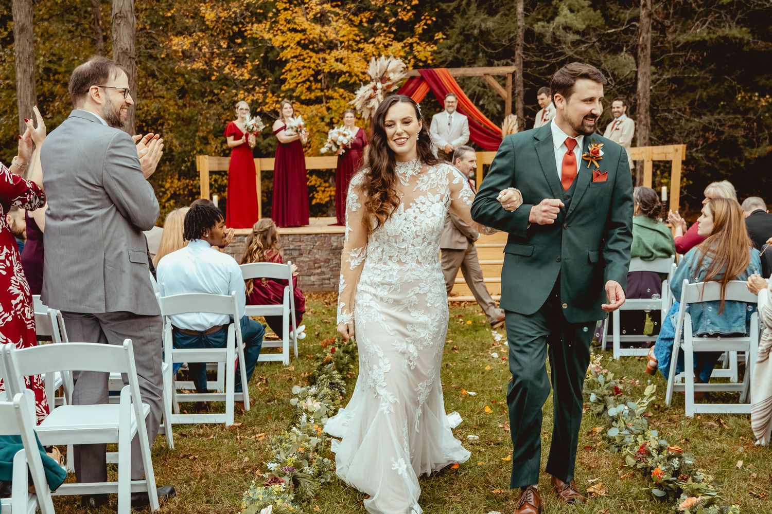 Bride and groom walk hand-in-hand down outdoor aisle after fall wedding ceremony surrounded by autumn foliage.