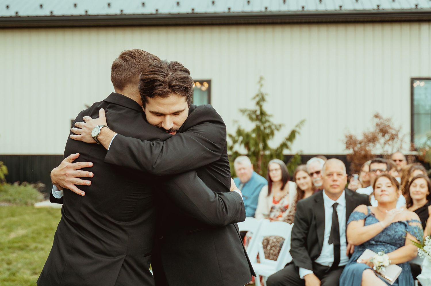 Two men in suits share an emotional hug at an outdoor wedding ceremony with guests seated in background.