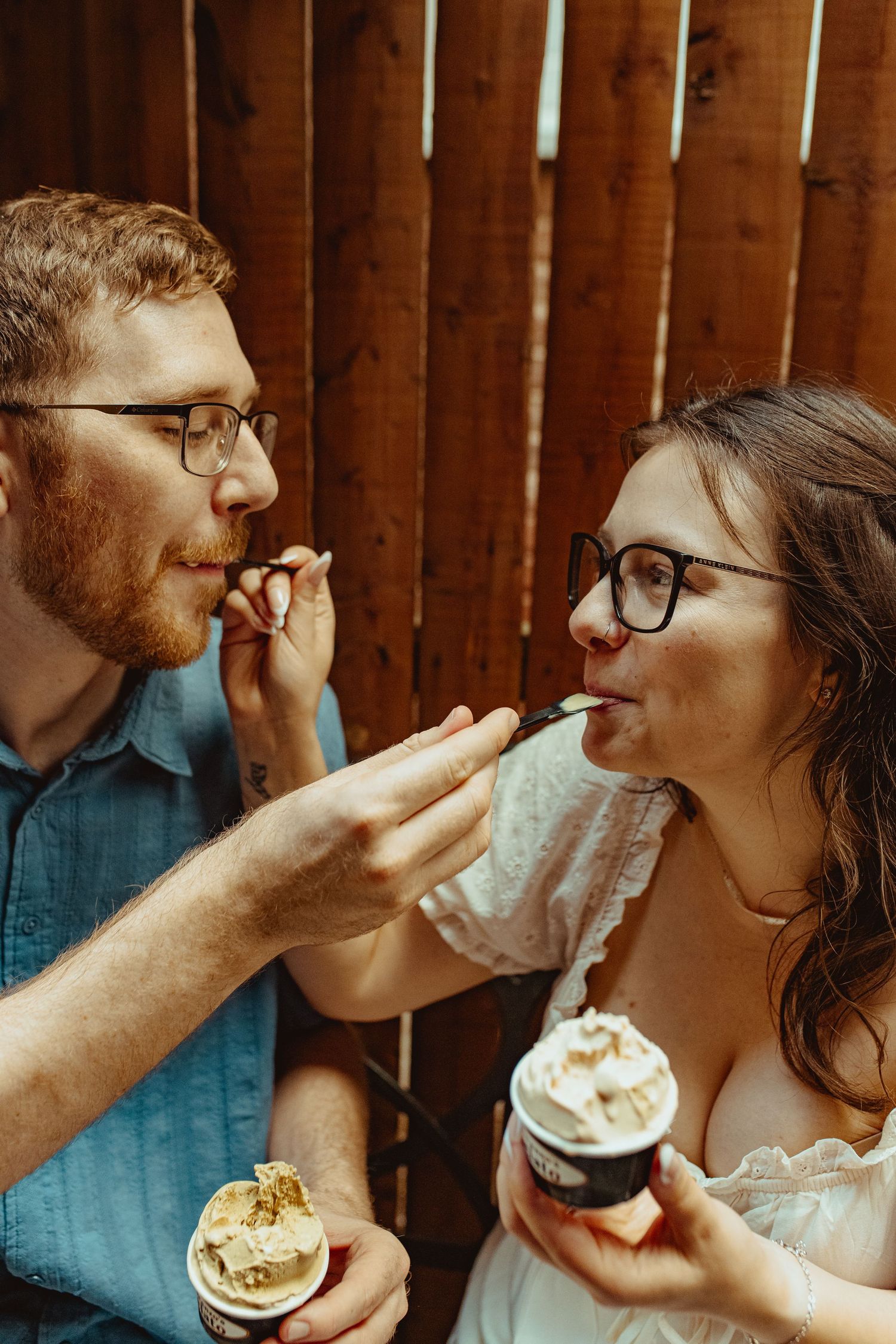 Romantic couple feeding each other ice cream, both wearing glasses, against a wooden background.