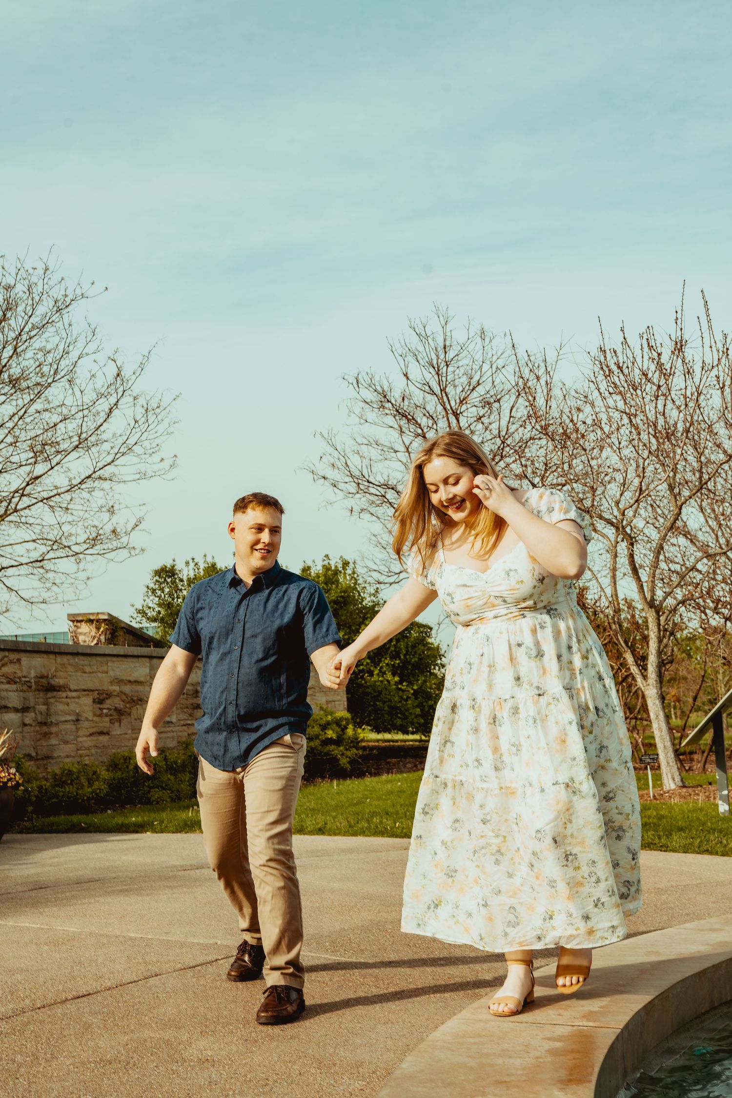 A couple holds hands and walks together outdoors, smiling, with bare trees and blue sky in the background.