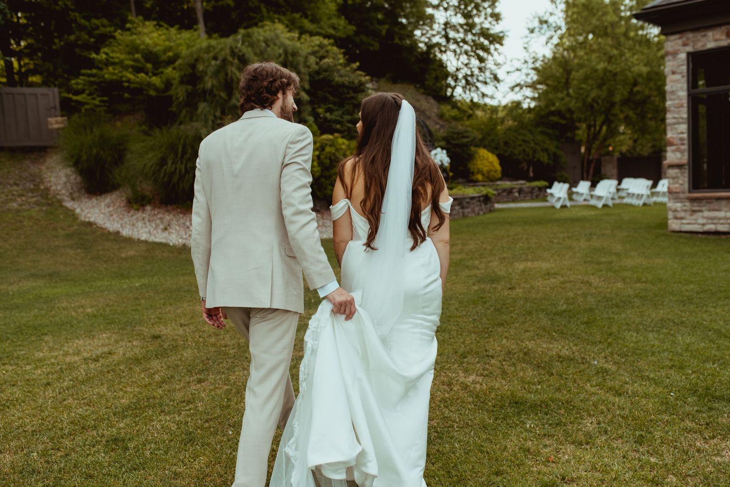 A bride and groom in matching white attire walk hand in hand across a lush green lawn.