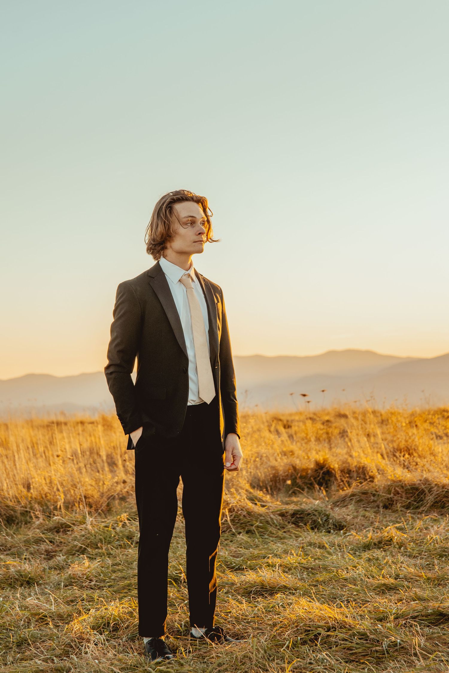 A suited figure stands in golden sunlit field with mountains visible in the background during sunset.